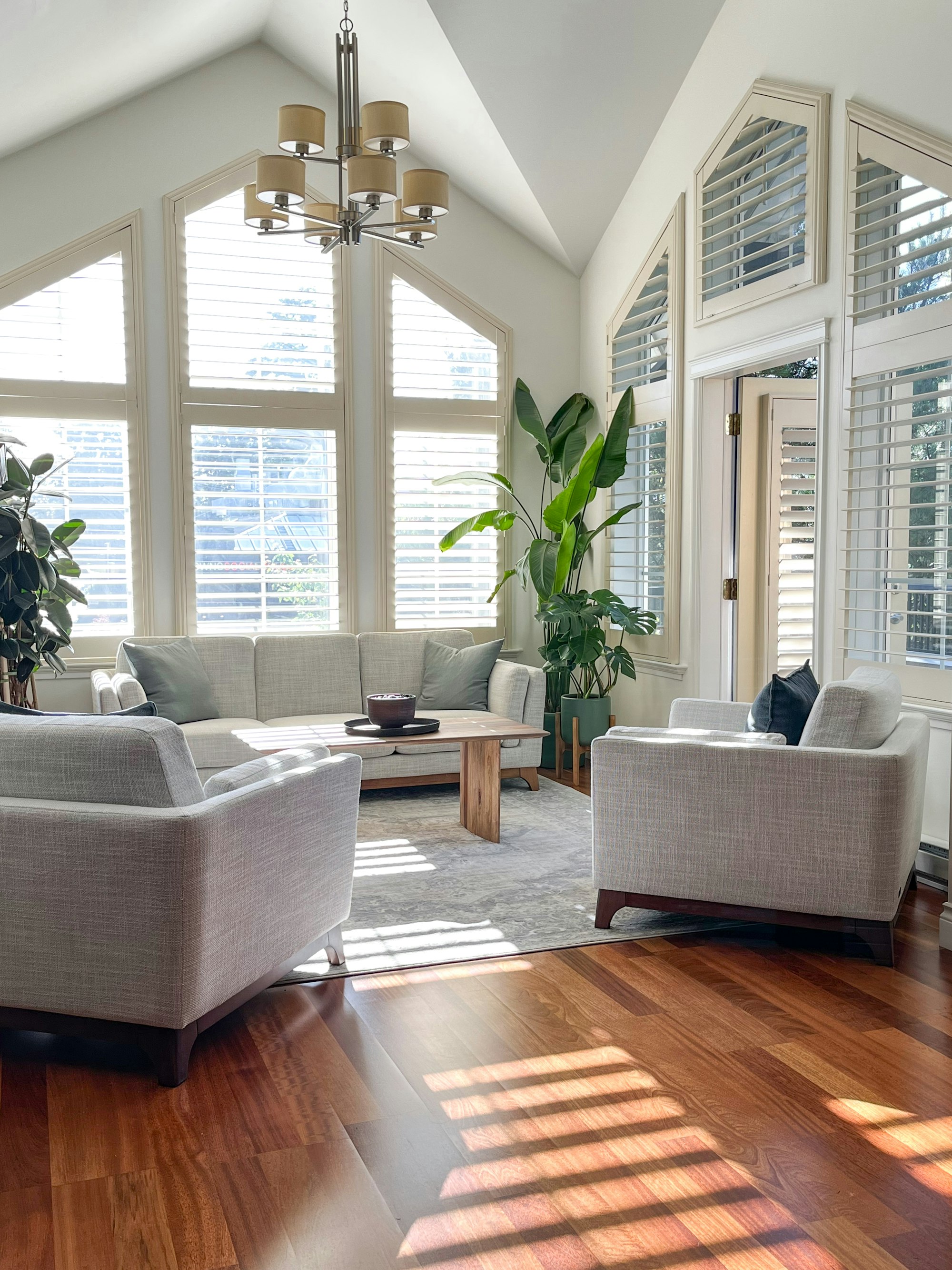 Bright living room with plantation shutters and shades, sunlight falling across warm wood floors